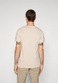 Mand vendt væk iført en beige T-shirt og khaki bukser, som viser farverige sommerfuglstattooer på begge overarme.
