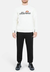 Sudadera blanca con logo "ellesse", pantalones deportivos negros y zapatillas deportivas grises. El conjunto presenta un corte holgado y un diseño sencillo.