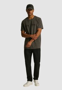 Szary T-shirt z czarnym logo, w połączeniu z czarnymi dżinsami i białymi trampkami. Model nosi również pasującą szaro-czarną czapkę. Gładkie tło.