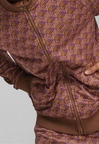 Veste à fermeture éclair marron avec un motif géométrique en violet et orange, en tissu texturé avec poignets et taille élastiqués.