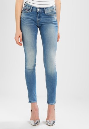 Femme portant un jean skinny bleu clair et des talons hauts argentés, debout devant un fond clair uni.