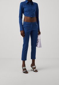 Jasnoniebieska skrócona bluzka, pasujące niebieskie spodnie z podwiniętymi mankietami, sandały z metalowym zapięciem oraz lawendowa torebka. Gładka faktura na całej powierzchni.