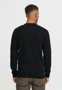 Pulover negru tricotat cu guler rotund și mâneci lungi. Materialul pare moale, cu un design croiți, asociat cu pantaloni texturați de culoare maro închis.