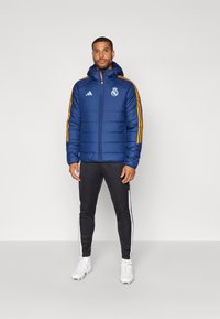 Giacca puffer blu con cappuccio, con strisce arancioni sulle maniche e un logo di squadra. Abbinata a leggings neri e scarpe bianche.