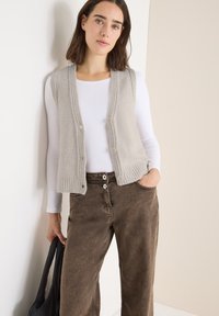 Hellgraue, knöpfbare Weste mit strukturiertem Rippenmuster, getragen über einem weißen langärmeligen Shirt. Braune weit geschnittene Hose mit zwei Knöpfen und einer Tasche.