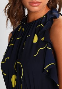 Navy mekko, jossa on keltaisia abstrakteja kuvioita, röyhelöhihoja ja laskostettu muotoilu. Silkkinen rakenne, tiukka vartalo ja laineileva kangas.