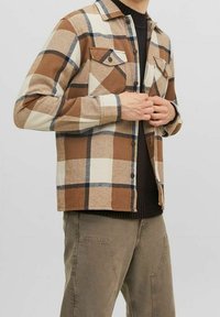 Bruin en crèmekleurig geruit shirt met puntig kraag, knoopsluiting aan de voorkant, twee borstzakken en textuurstof, gelaagd over een zwarte trui.