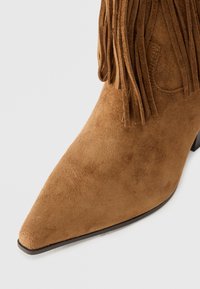 Botine din suede maro, cu vârful ascuțit, având franjuri în partea superioară și un toc mic, conic. Textură netedă cu detalii minime.