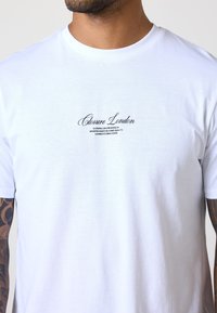 Hvid bomulds T-shirt med rund halsudskæring og korte ærmer. Teksten foran lyder "Closure London" i en elegant sort skrift.