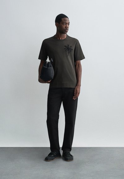 Tricou din bumbac de culoare verde măsliniu închis, cu broderie neagră sub formă de floare pe partea stângă a pieptului, purtat cu pantaloni din denim negri și adidași negri.