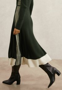 Robe mi-longue en maille côtelée vert foncé avec détail d'ourlet blanc et rayures verticales. Associée à des bottines noires à bout pointu et talons.