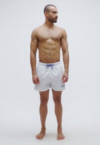 Fehér fürdőnadrág kék húzózsinórral, bal lábán kis logóval. A szövet könnyűnek és simának tűnik. Meztelen lábú modell.