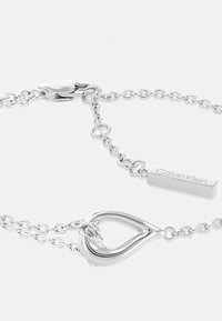 Zilveren kettingarmband met een hartvormige schakel en een rechthoekig hangtag gegraveerd met "Calvin Klein." Glad, gepolijst afwerking.