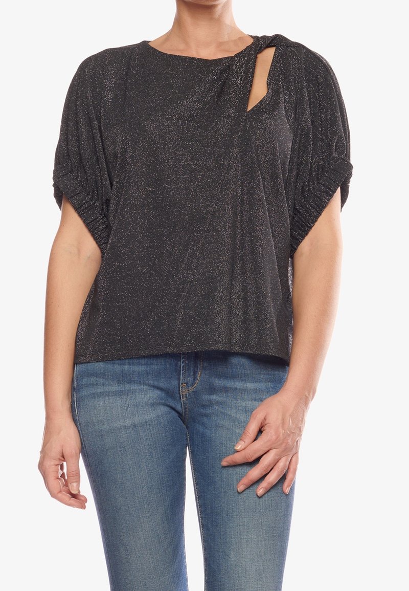 Haut noir texturé avec manches courtes bouffantes, présentant un détail noué et une découpe latérale, fabriqué dans un tissu scintillant. Porté avec un jean bleu.
