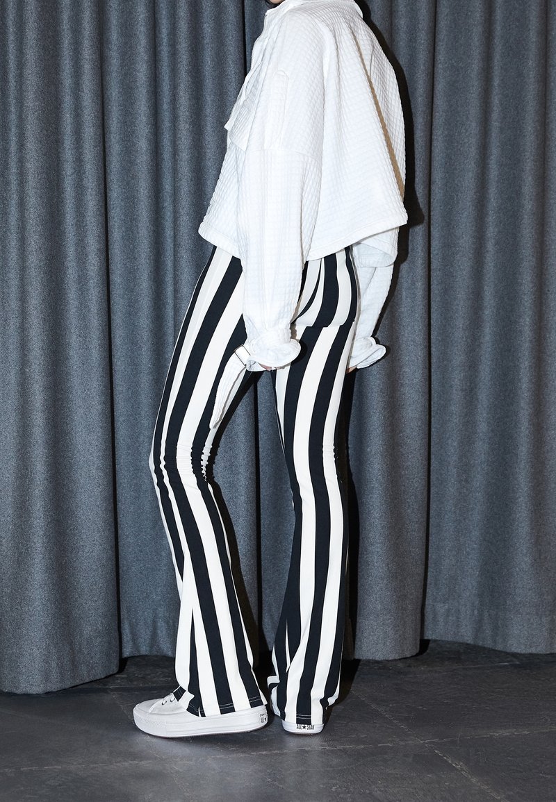 Black and white striped flared pants with a snug fit. Worn with a loose white textured top and white sneakers. Background is gray.