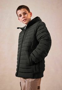 Giacca nera imbottita con cappuccio, dotata di una chiusura con zip, orlo a coste e texture trapuntata e gonfia. Indossata sopra un top grigio, su uno sfondo semplice.