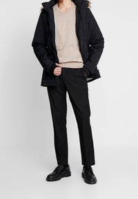 Schwarze Parka mit Kapuze, beiger V-Ausschnitt-Pullover, schwarze Hose und schwarze Schuhe. Der Mantel hat einen Fellbesatz. Glatte Texturen durchgehend.