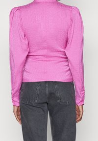 Vaaleanpunainen pitkähiha T-paita, jossa on teksturoitu kuvio; siinä on vartalonmyötäinen malli ja röyhelöhihat. Päällä tummanharmaat korkeavyötäröiset farkut.