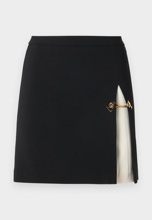 Crna mini suknja s bočnim prorezom koji otkriva krem podstavu, sadrži zlatne lančane detalje i elegantnu, glatku teksturu tkanine.