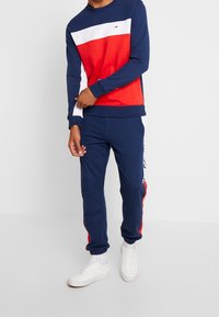 Mörkblå sweatshirt med röda och vita ränder, kombinerad med matchande mörkblå joggers med röda sidodetaljer. Vita sneakers fulländar outfiten.