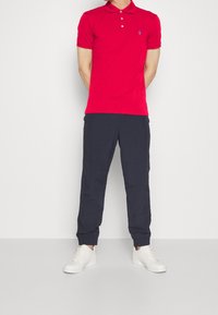 Červené polo tričko s límečkem a třemi knoflíky, doplněné o tmavě modré teplákové kalhoty. Outfit je dokončen bílými teniskami.