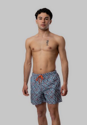 Uomo senza maglietta in piedi, indossa pantaloncini da bagno con fantasia e coulisse arancione, espressione neutra, tatuaggio con caratteri coreani sulla parte superiore del petto.