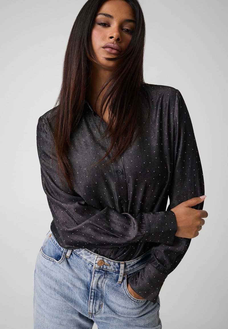 Femme aux cheveux longs et lisses porte une chemise noire à pois à manches longues rentrée dans un jean bleu clair, posant une main sur la hanche.