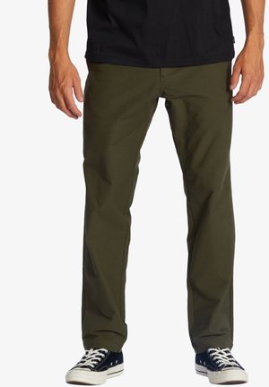 Unterkörper einer Person, die olivgrüne Hose, ein schwarzes Hemd und schwarz-weiße Turnschuhe vor einem weißen Hintergrund trägt.