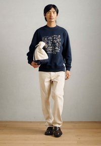 Ναυτικό φούτερ με γραφικό τύπωμα, κρεμ παντελόνι και σκούρα παπούτσια; κρατώντας μια τσάντα από ύφασμα δύο τόνων με καμπύλο σχήμα. Απλός και χαλαρός σχεδιασμός.