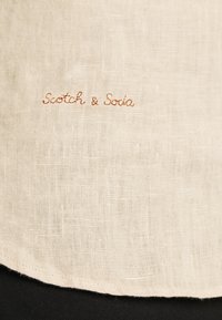 Tessuto di lino color crema con il testo ricamato "Scotch & Soda" in color ruggine, caratterizzato da una texture liscia e un orlo curvo.