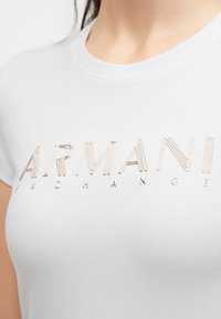 Κοντινό πλάνο λευκού T-shirt με στρογγυλή λαιμόκοψη και μεταλλικά χρυσά γράμματα "ARMANI EXCHANGE" στο μπροστινό μέρος.