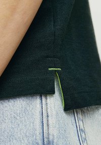 Chemise vert foncé en tissu texturé, avec un accent de couture vert clair à l'ourlet, associée à un denim bleu clair.