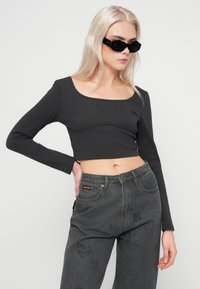 Ξανθιά γυναίκα που φοράει μαύρο κοντό τοπ με μακρύ μανίκι, σκούρες γκρίζες τζιν σε ψηλή μέση με διακριτικά σχέδια, και μαύρα ορθογώνια γυαλιά ηλίου μπροστά σε λευκό φόντο.