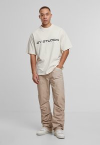 Nuori mies seisoo yksi käsi taskussa, päällä valkoinen "2Y STUDIOS" -t-paita, beige cargo-housut ja valkoiset tennarit yksinkertaista taustaa vasten.