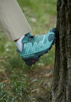 Pied chaussé d'une chaussure de trail Salomon bleu-vert posée sur une écorce d'arbre en extérieur, pantalon beige et chaussette blanche visibles.