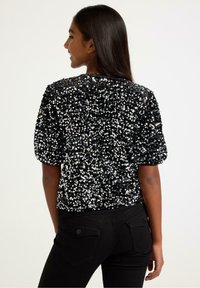 Černý top zdobený flitry s kulatým výstřihem a krátkými nafouklými rukávy, s přiléhavým střihem. Kombinováno s černými džínami z denimu.