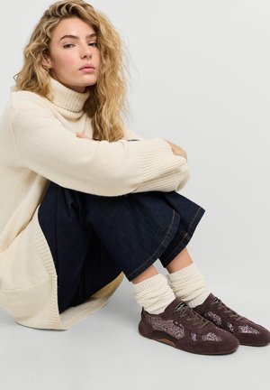 Giovane donna con capelli biondi ricci, che indossa un maglione dolcevita color crema, pantaloni scuri, calzini bianchi e scarpe da ginnastica marroni glitterate, seduta con le ginocchia alzate.