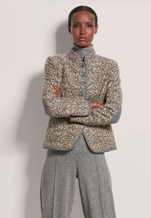 Tan patterned wool jacket with gray accents, five front buttons, and high collar. Worn over a gray turtleneck and houndstooth trousers.