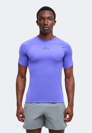 Egy férfi áll, előre néz, visel egy testhezálló, rövid ujjú, lila Nike Pro pólót és világosszürke Nike rövidnadrágot, semleges háttér előtt.