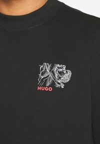 Maglietta nera a maniche lunghe con un design testurizzato, elementi grafici bianchi e un logo rosso "HUGO" sul davanti.