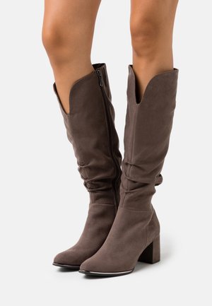 Bottes hautes jusqu'au genou en daim marron avec un design souple, un bout pointu et un talon bloc. Elles présentent une fermeture éclair latérale et une découpe en V distinctive en haut.