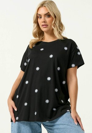 Blondynka w luźnej czarnej koszulce z rozproszonymi małymi białymi nadrukami stokrotek, w połączeniu z jasnoniebieskimi dżinsami, stoi na jednolitym tle.