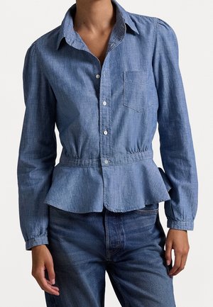 Kobieta w dopasowanej dżinsowej koszuli z guzikami, kieszenią na piersi, talią z baskinką oraz długimi rękawami, w połączeniu z ciemnymi dżinsami.