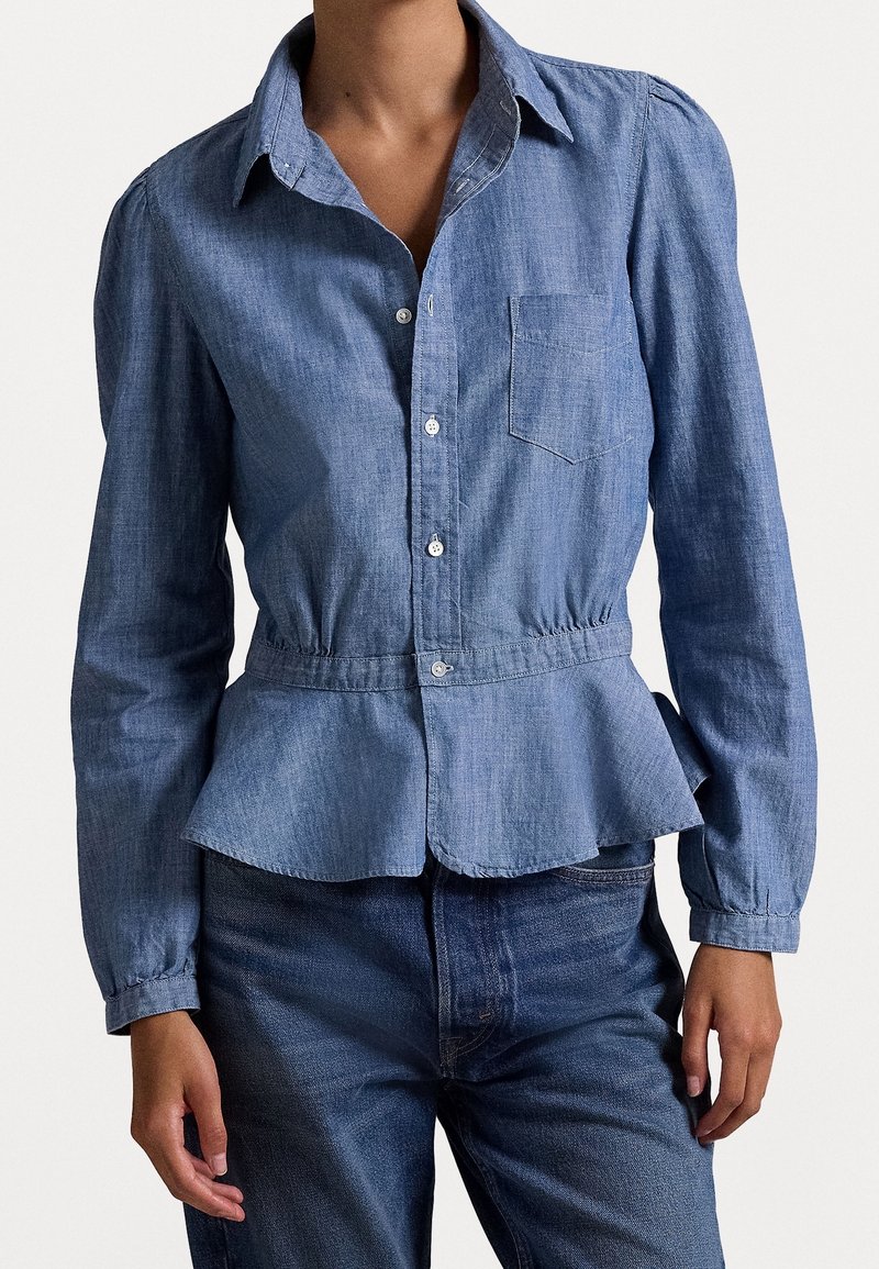 Femme portant une chemise en denim ajustée avec des boutons, une poche poitrine, une taille peplum et des manches longues, associée à un jean denim foncé.