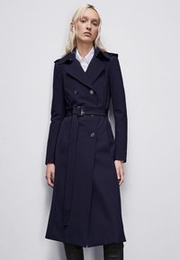 Cappotto trench blu navy con design doppiopetto, vita cintata e spalline, realizzato in tessuto morbido con una vestibilità su misura.