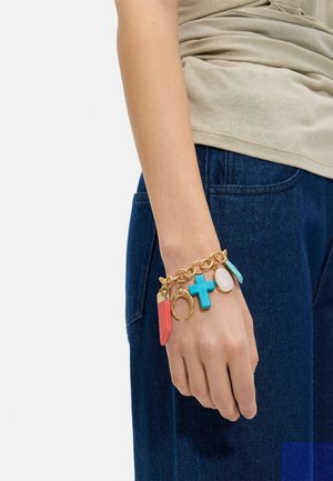 Złota bransoletka z łańcuszkiem z kolorowymi zawieszkami, w tym turkusowym krzyżem, koralowym wisiorkiem, podkową i owalnymi kamieniami na nadgarstku osoby.