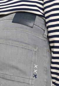 Los vaqueros de mezclilla gris tienen un acabado texturizado, un parche de cuero azul con un logotipo en relieve y costuras decorativas en blanco y azul. Parte de una camiseta a rayas es visible.