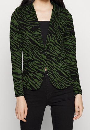 Woman wearing a fitted green and black zebra-striped blazer with black buttons over a black top and black pants, standing with arms relaxed.