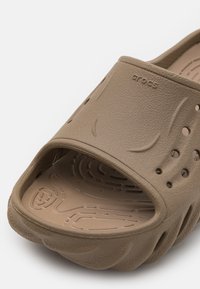Brown, rubber slide sandal featuring an open toe, texture pattern, ventilation holes, and a contoured footbed. Branding visible on the top.