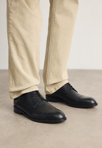 Chaussures habillées en cuir noir avec une finition lisse, bout rond, design à lacets et semelle en caoutchouc plate, assorties à un pantalon beige clair.